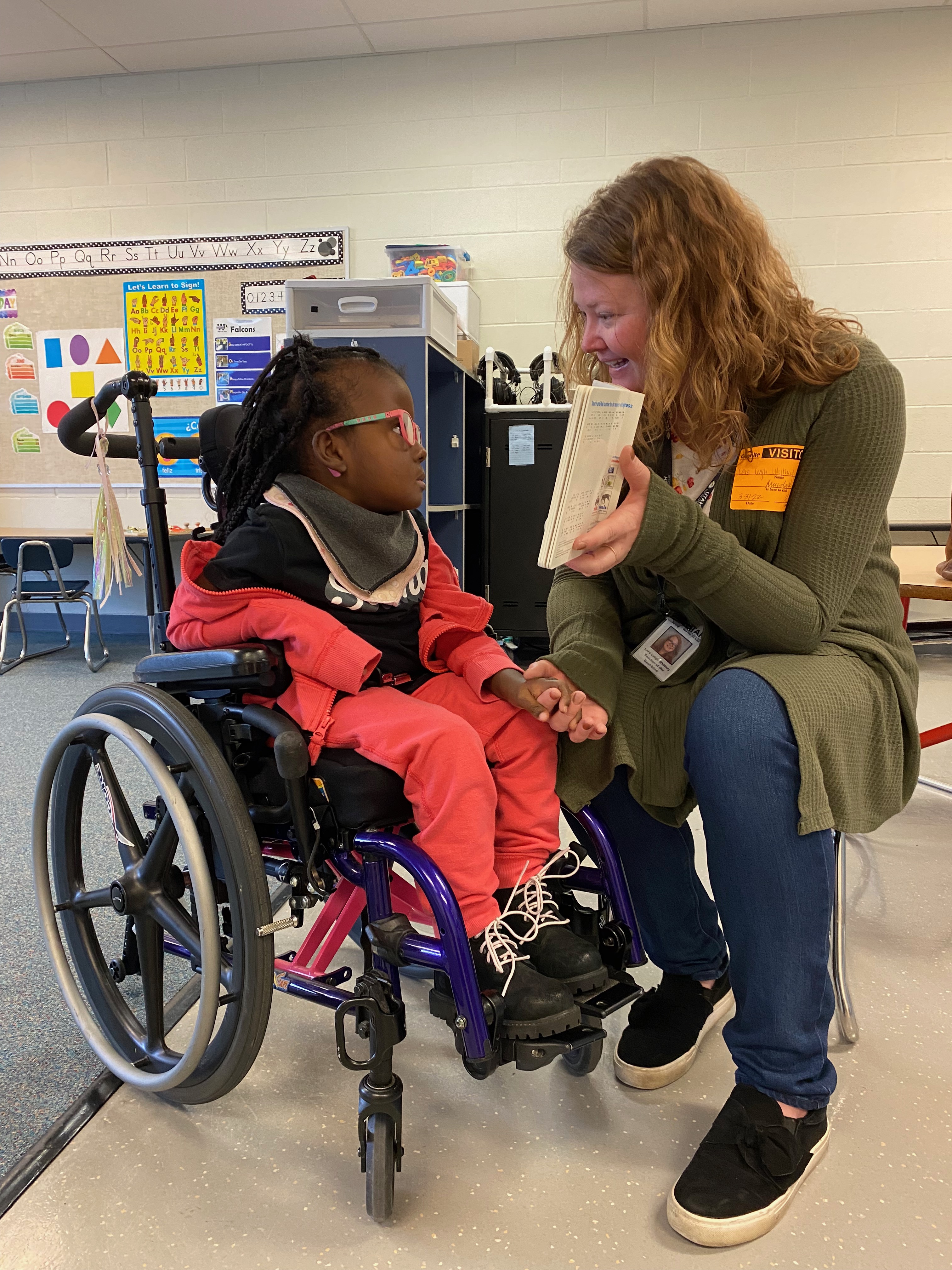 A young female student who is deaf-blind sits in a wheelchair. Her teacher sits beside her holding a braille book. The student looks at the book and holds her teacher’s hand.