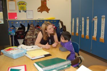 An adult woman, an educator, and a student who is deaf-blind, sit at a table in a classroom. He has his hand on her arm. They appear to be having a conversation.