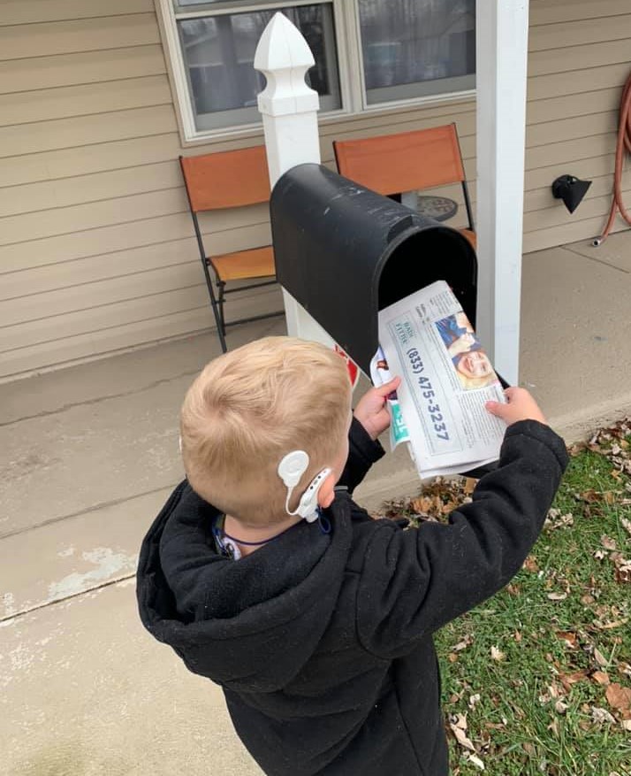 Un niño está sacando un periódico de un buzón. Tiene un implante coclear.