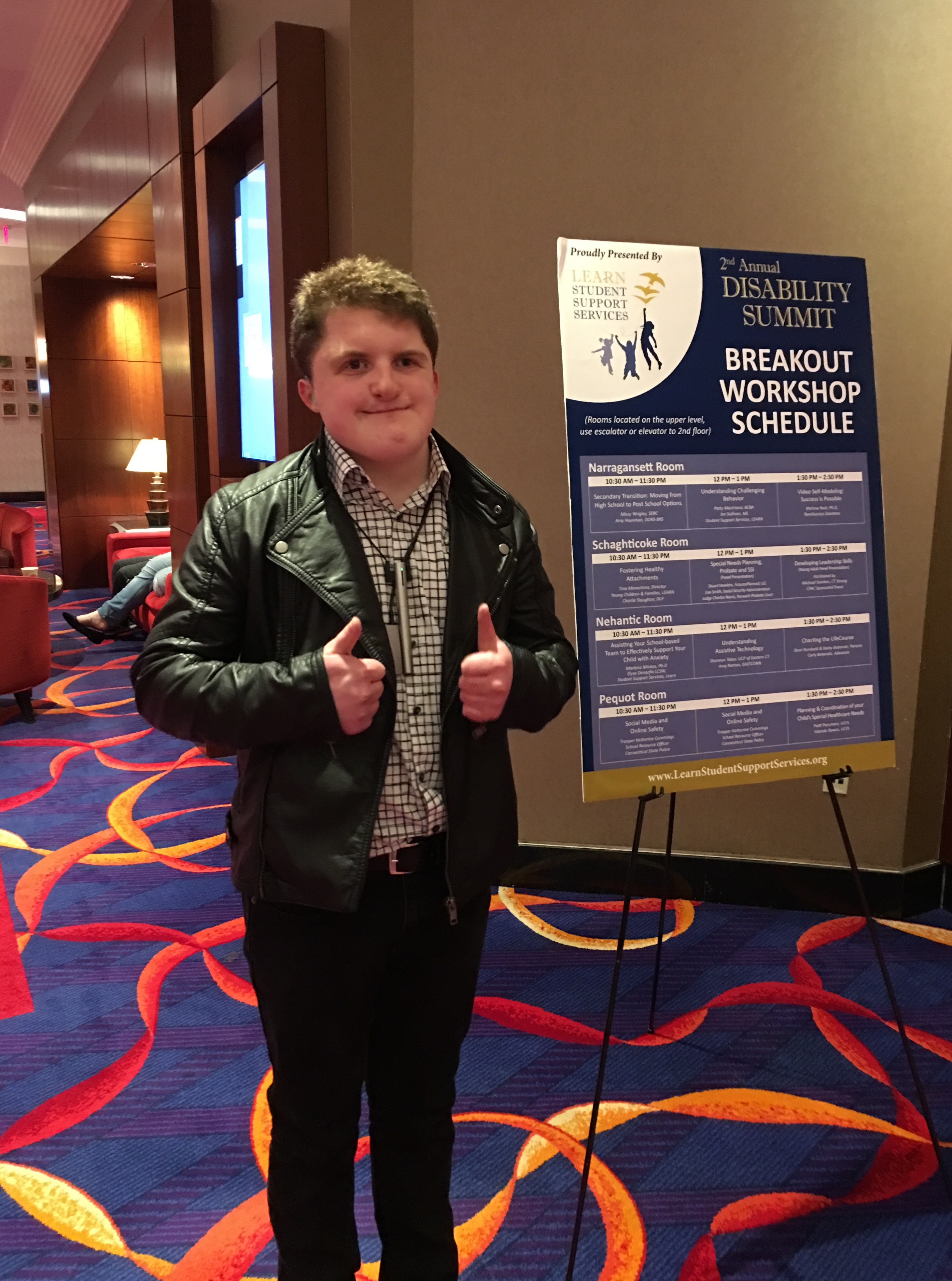 Alex in front of poster at conference making the thumbs up gesture.
