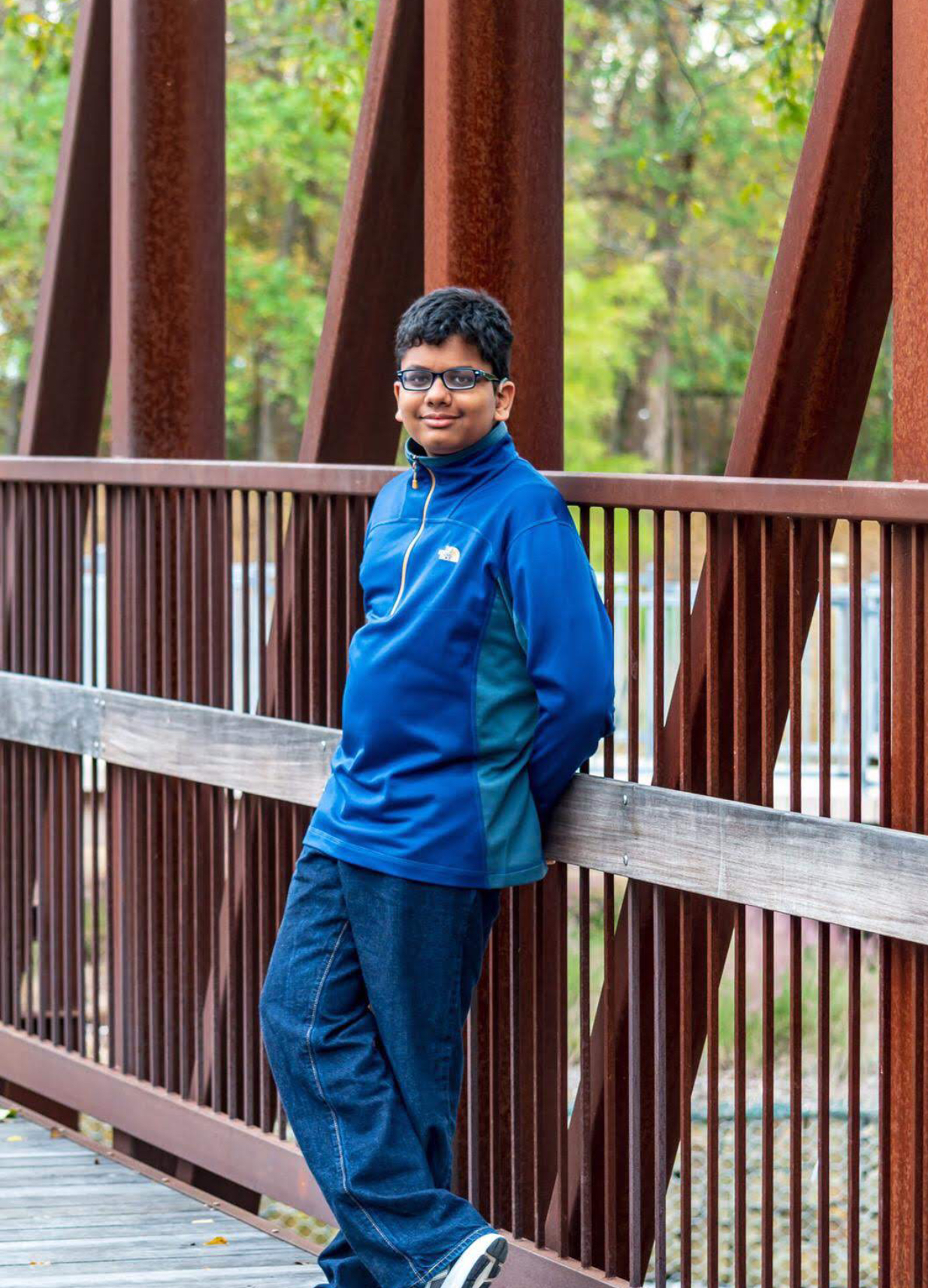 A 14-year-old boy with Usher Syndrome is leaning against the railing of a  bridge smiling at the camera. He looks relaxed and happy.