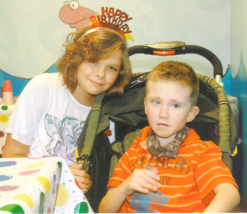 A young boy wearing an orange shirt is sitting in a stroller with a young girl standing next to him. The young girl has a "Happy Birthday: headband on her head.