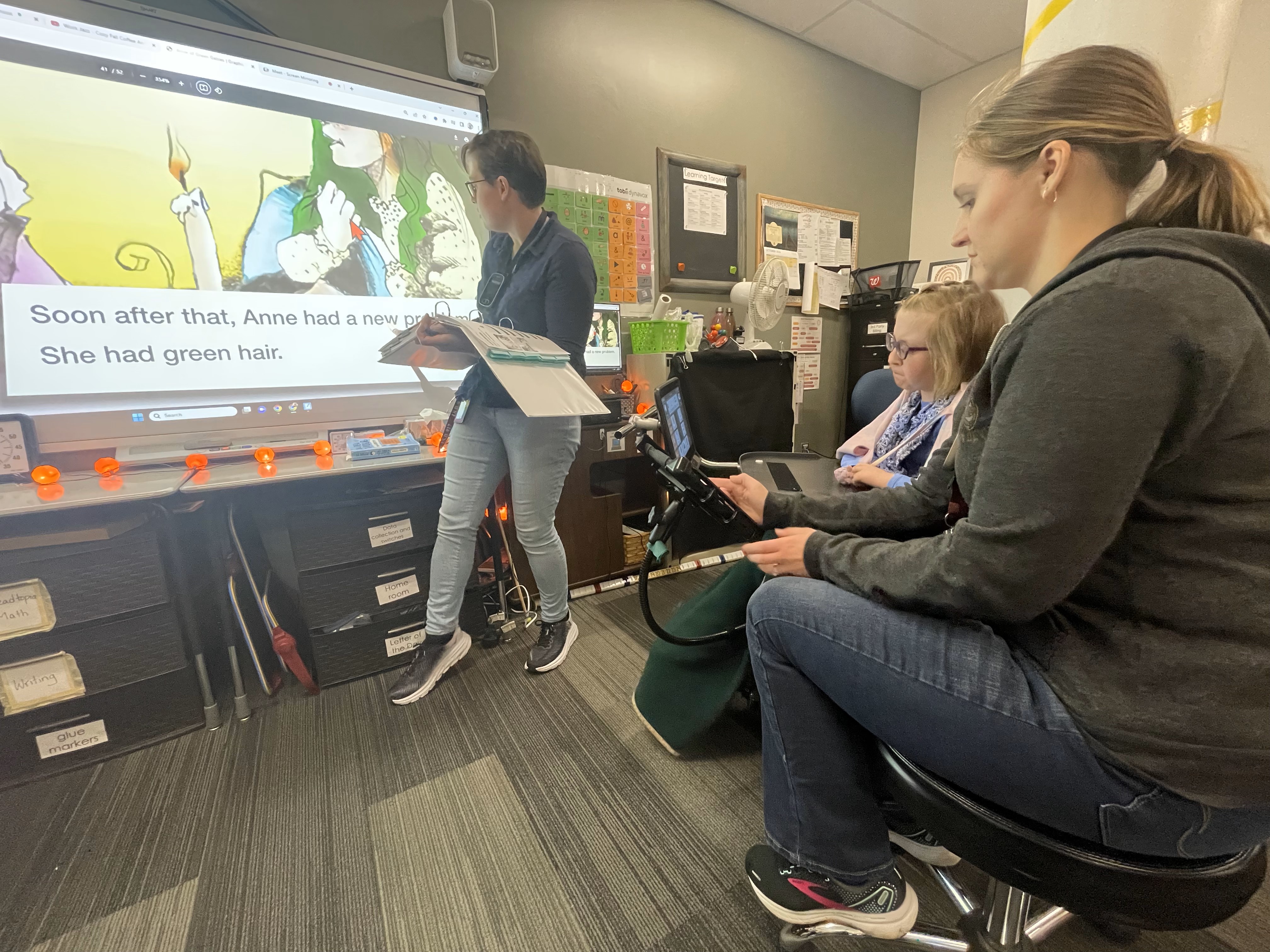 A young teenage girl works with her teacher and her intervener in a classroom. She uses an AT device to follow the reading lesson.