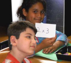 An adolescent boy with deaf-blindness sits at desk with a girl who is his classmate. She is holding a piece of paper that says 2 x 3.