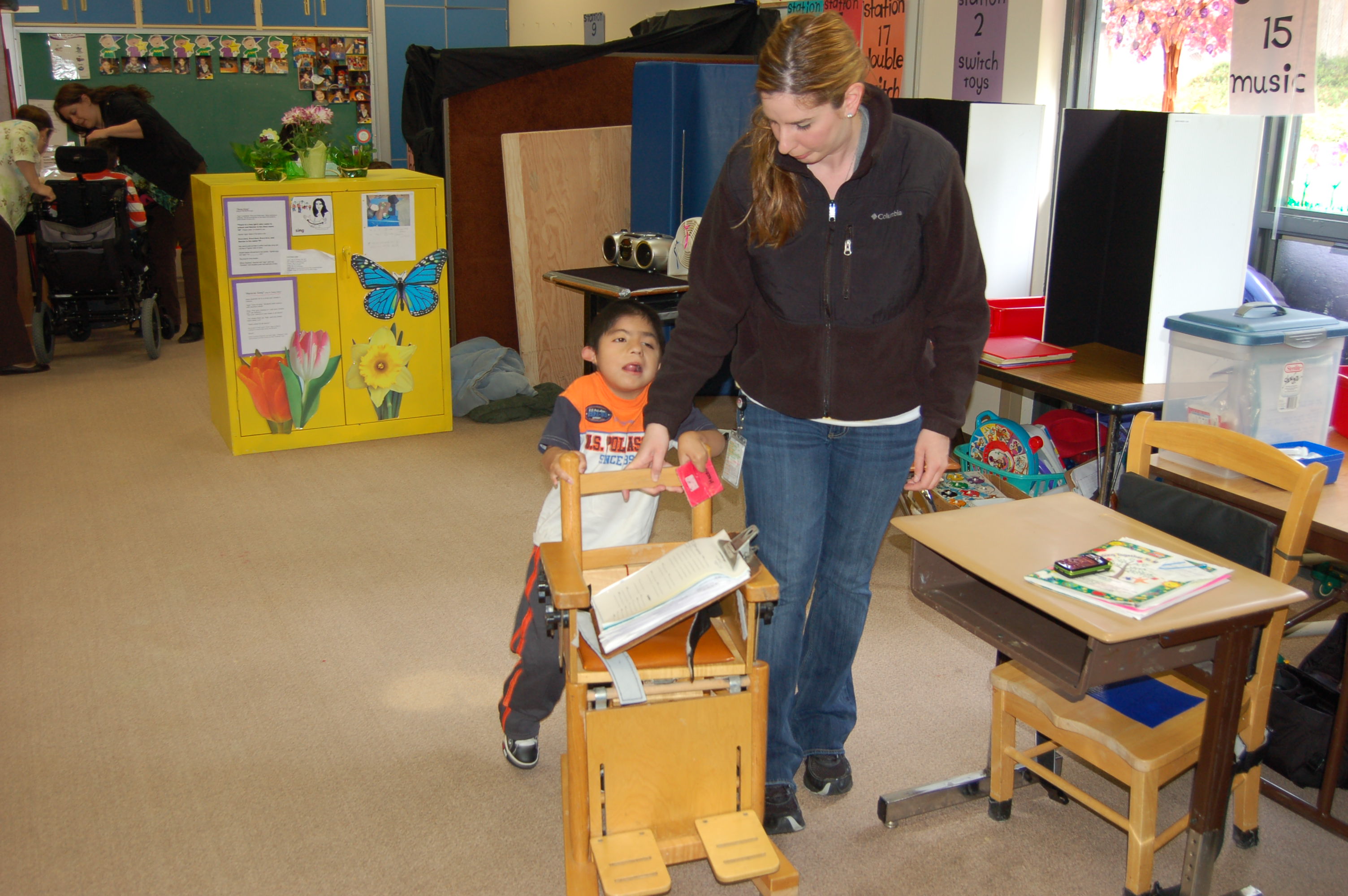 A young boy in a classroom is holding onto a specialized chair to support him while walking. He is assisted by an adult who also has her hand on the back of the chair.
