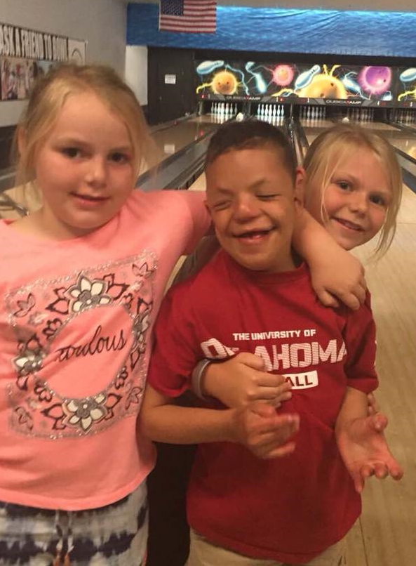 A boy who is deaf-blind is at a bowling alley with two girls. They have their arms around each other and are smiling.