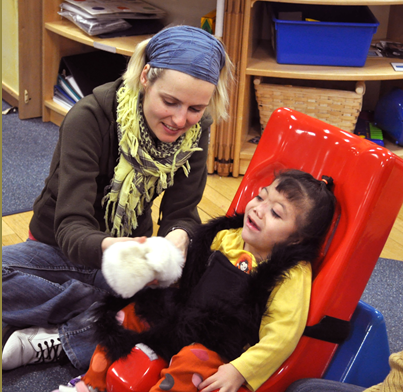 An interveners interacting with a child in a booster chair.