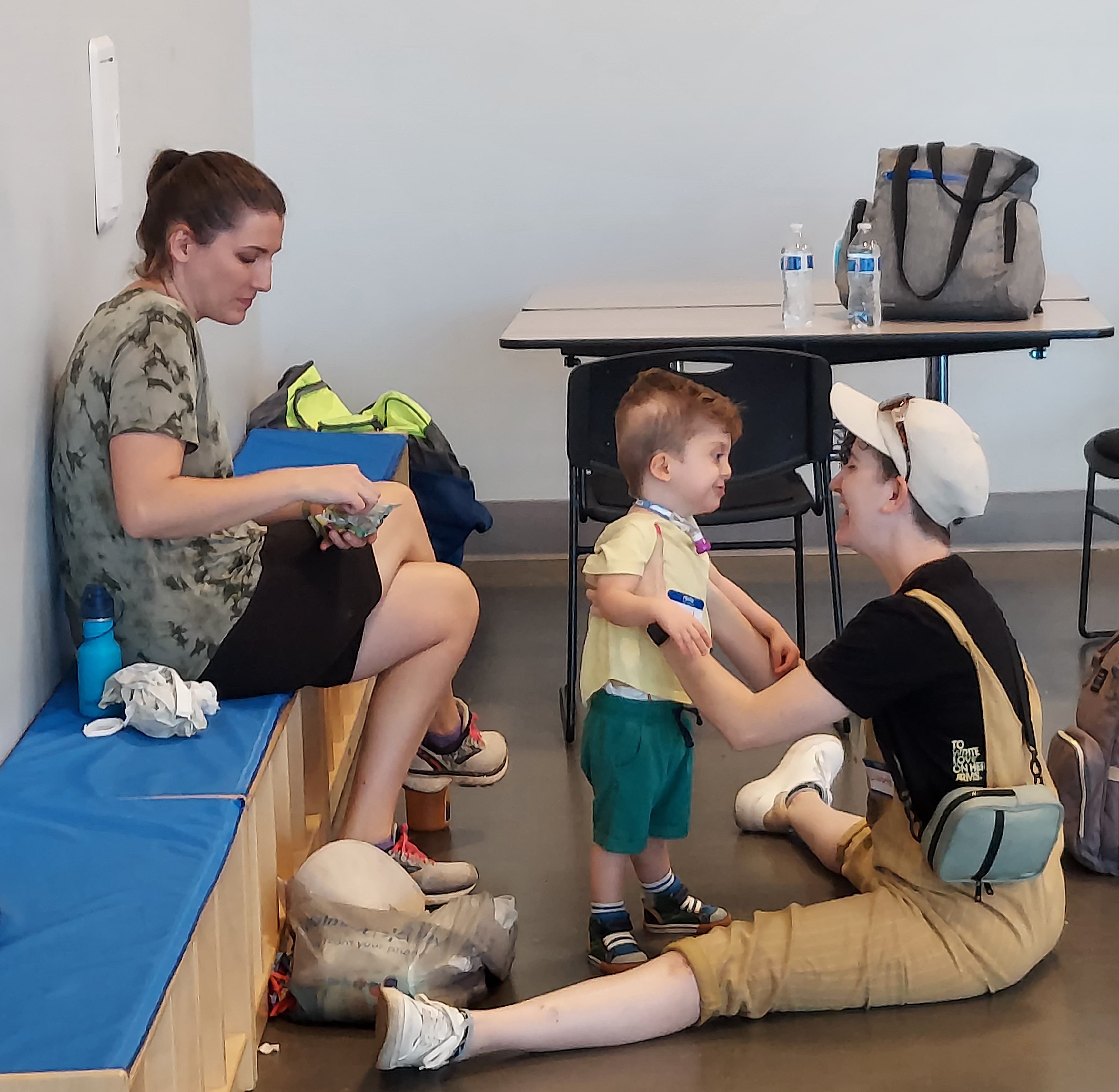 A young boy who is deafblind stands in a classroom. His teacher sits on the floor in front of him. She supports him by holding his arms. His mother sits on the bench behind them.