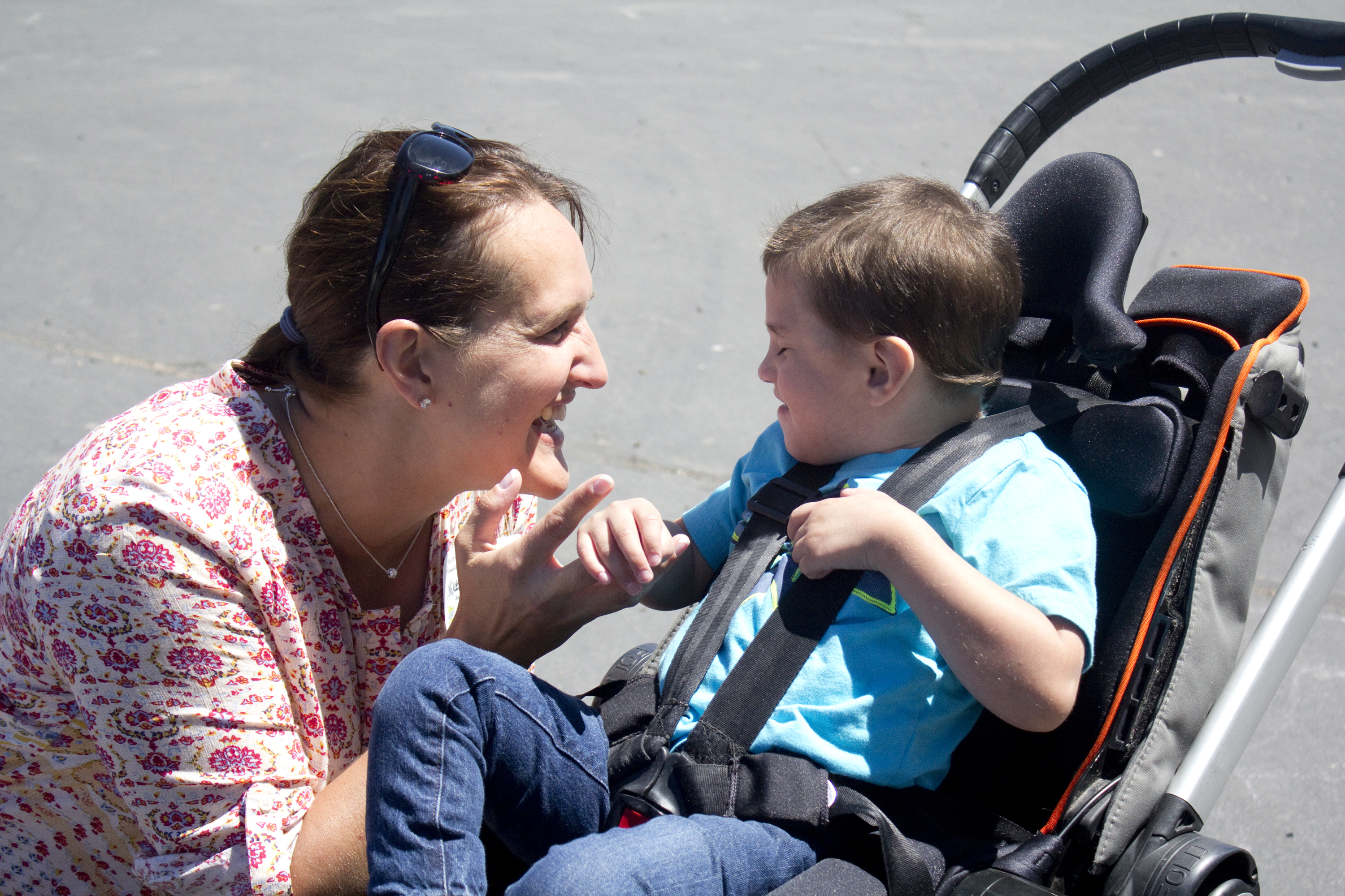 A young boy who is deafblind sits in his stroller. His teacher kneels beside him and touches his hand. They both smile