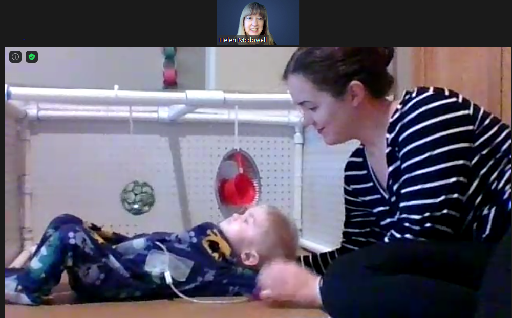 Screenshot of zoom call of teacher and child with parent. 9-month-old child lying on back with toys near by. His mom his learning over him, smiling gently.