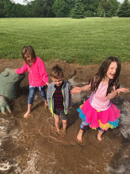 There are three children holding hands barefoot in a puddle.  There is a young girl on the left in a pink jacket and jeans. There is a young boy in the middle wearing a grey jacket, and another young girl on the right wearing a colorful tutu .
