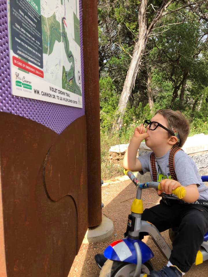 A young boy sitting on a bike, sucking his thumb, and looking at a nature sign.