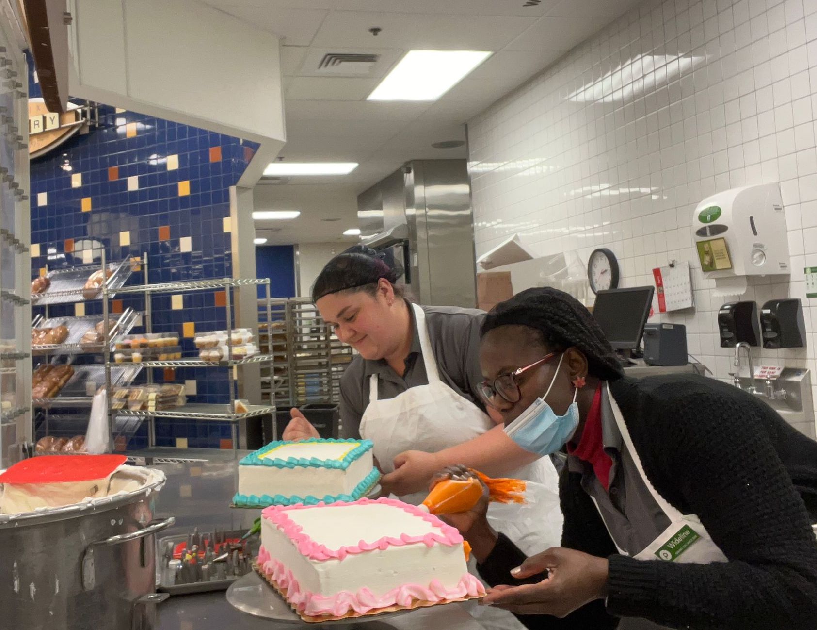 Two women decorating cakes with icing bags in a commercial kitchen.