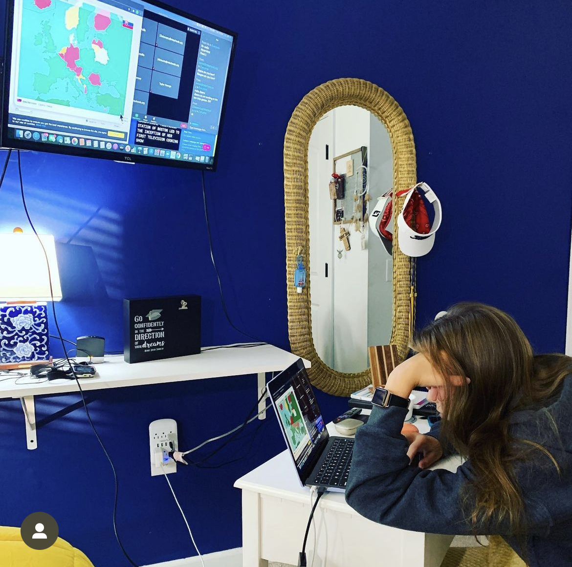 A teenage girl works on a laptop. There is a large monitor screen on the wall in front of her.