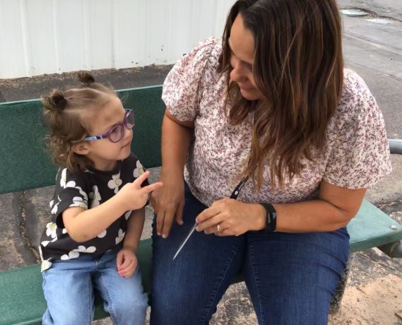 A young girl who is deafblind sits beside an adult female on an outdoor bench. The adult watches as the child signs the number 2.