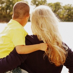 A mom in a black shirt with light hair is holding a young boy with a yellow shirt. They are facing away from the camera