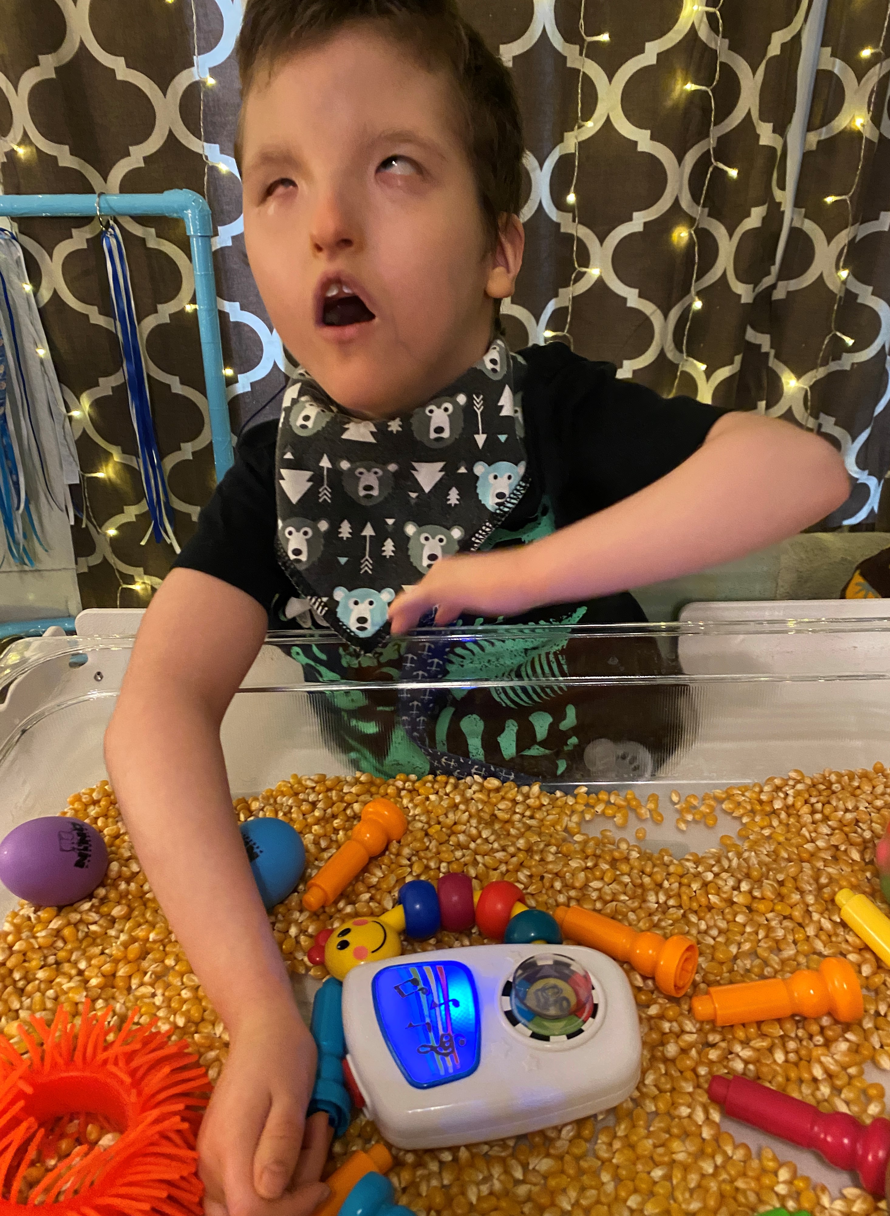 A boy with a visual impairment, about 8 years old, handling colorful toys in a clear box. The box also contains popcorn kernels.