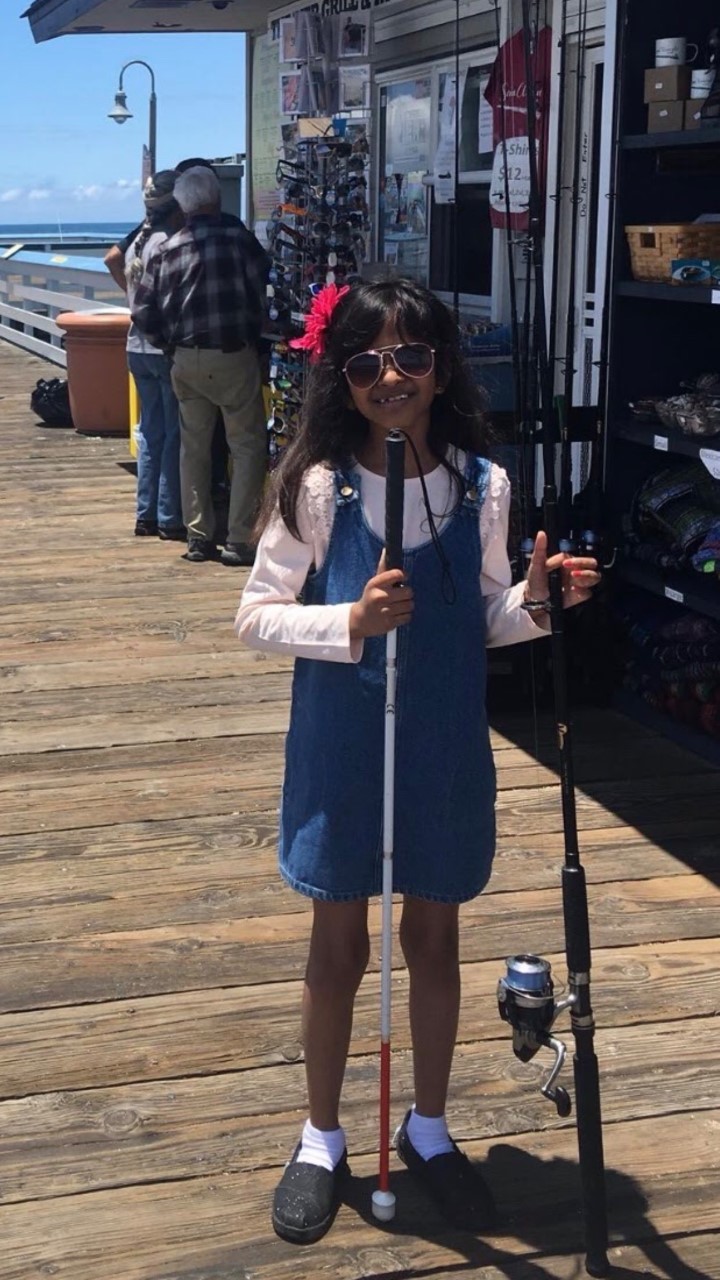 A girl standing on a dock. She is holding a white cane in her right hand and a fishing pole in her left.