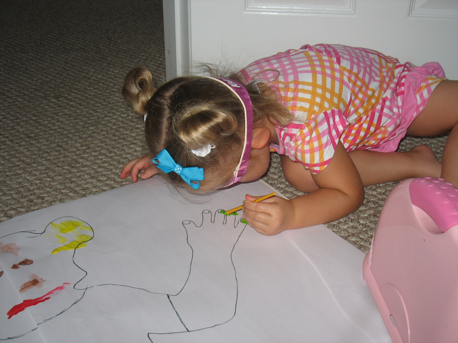A young girl in pigtails is sitting on the floor drawing a life-sized picture of herself on a very large piece of paper.