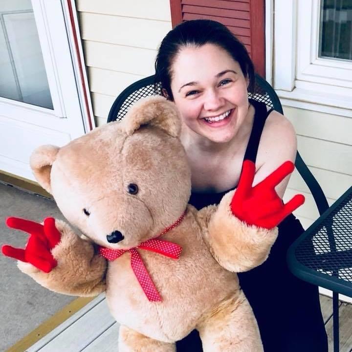 A smiling woman, sitting outside. She’s holding a large teddy bear who’s paws are in the shape of the ASL sign for “I love you.”