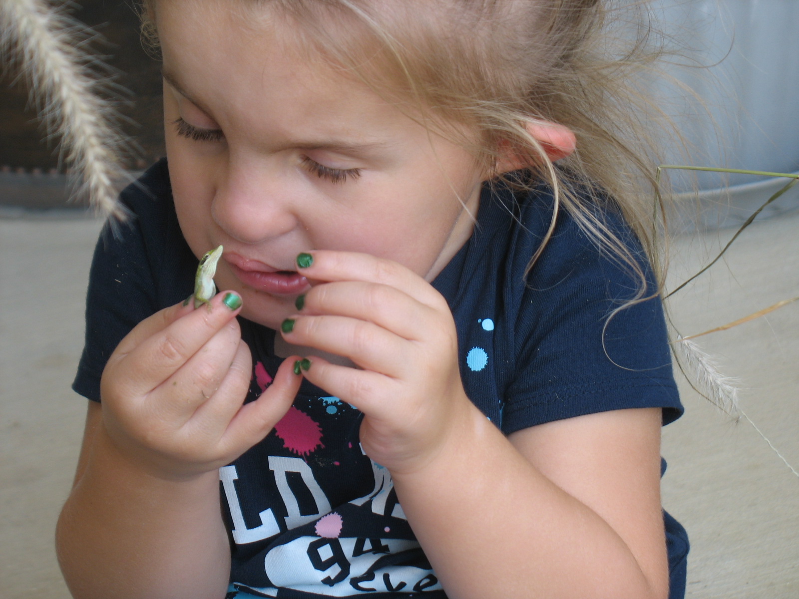 A young girl (approximately 5 years old) is holding a tiny lizard near her left eye to look at it carefully.