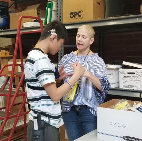 A student in his late teens who is deaf-blind is standing with an adult woman in a room with shelves and boxes. They are facing each other and communicating via tactile sign language.