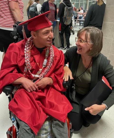 A young adult male student who is deaf-blind sits in a wheelchair. He is wearing a high-school graduation gown. His teacher kneels beside him. They are both smiling.