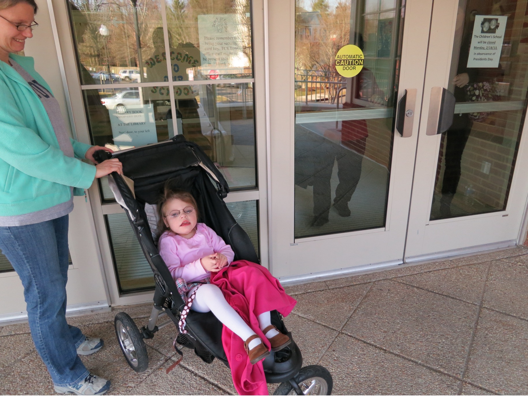 A young girl who is deafblind sits in a stroller outside. Her mom stands beside her.