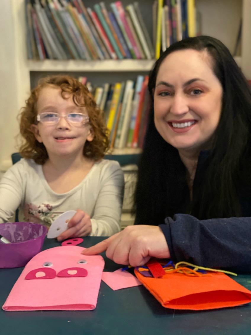 A mother and her 7-year-old daughter sitting side-by-side at a table working on a craft project. There are bookshelves behind them.