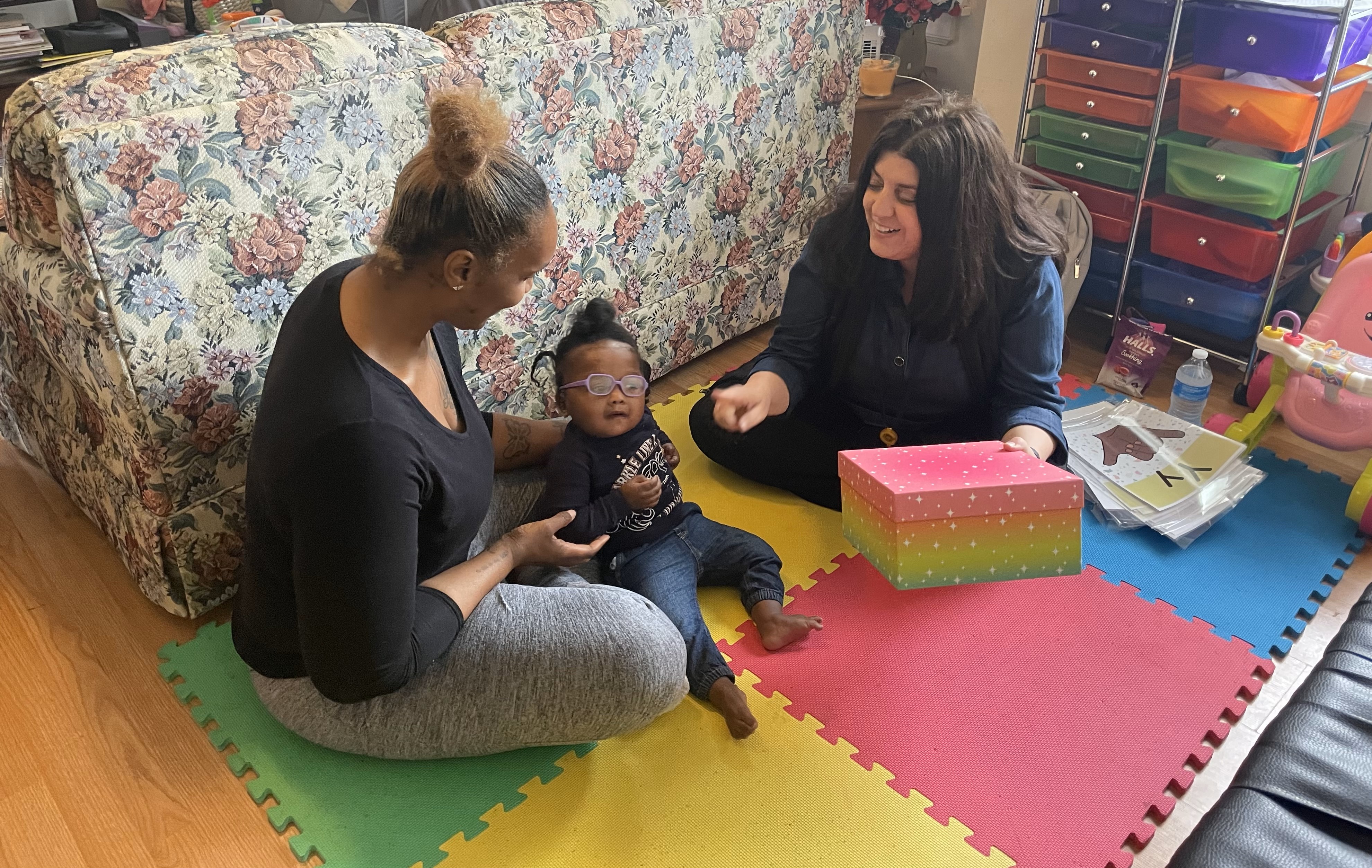 A baby who is deafblind sits on her mother's lap. A female EI provider sits near by.