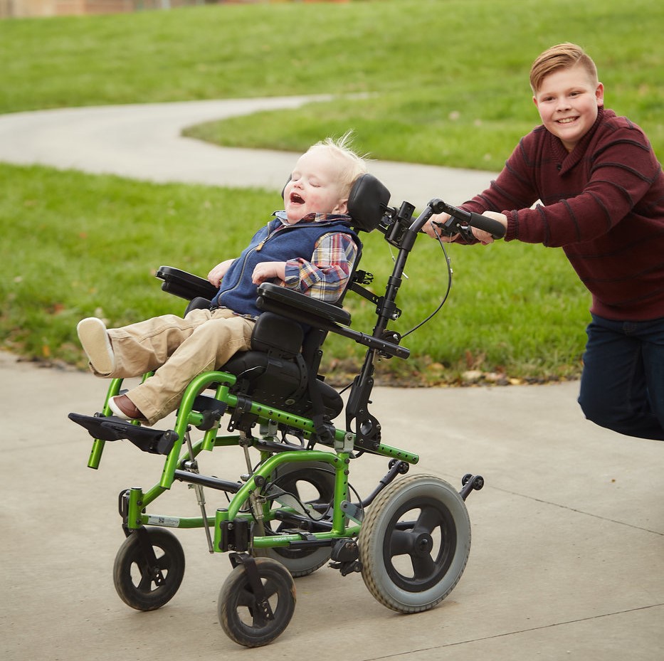 A 4-year old boy with deaf-blindness who is in a wheelchair is being pushed by his 9-year-old brother. It looks like they are going fast and both have big smiles.