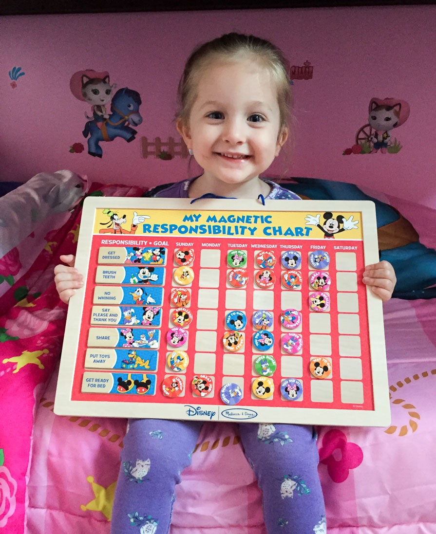 A young girl holding a chart containing Disney character magnets. It reads "My Magnetic Responsibility Chart.