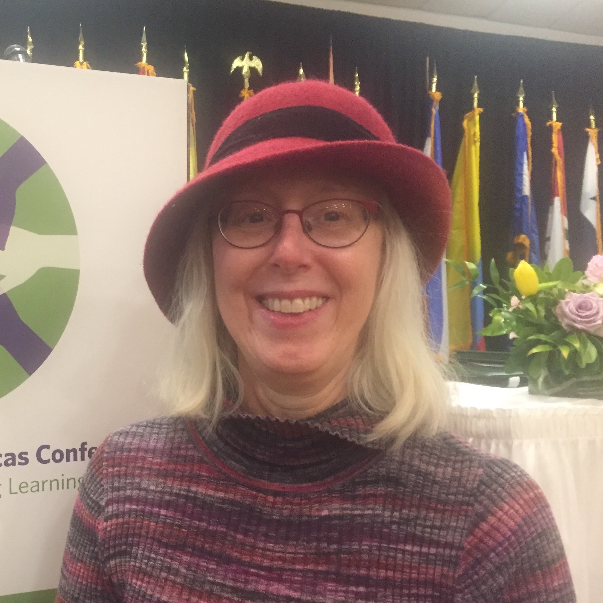 Michelle at a conference wearing a hat with several flags behind her, next to a conference poster.