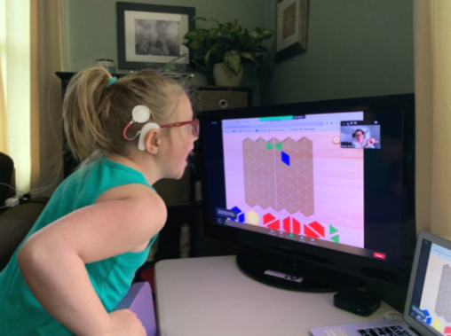 Young girl sitting at a desk looking with excitement at a computer screen. She is wearing glasses and has a cochlear implant.