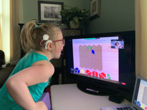 Chica joven está sentada en un escritorio y mira con emoción la pantalla de una computadora. Lleva lentes y un implante coclear.