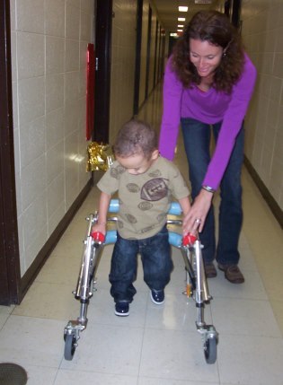 A young boy walking down hallway with walker.