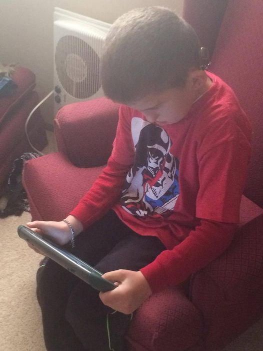 A young boy is sitting in a chair wearing a red shirt, he is looking at an ipad