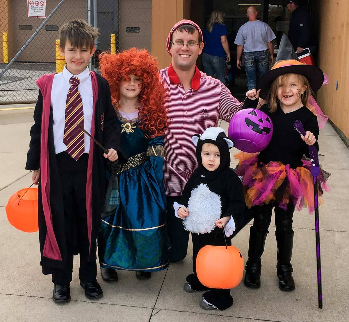 Teed family children (with their father) in Halloween costumes ranging from Harry Potter to an animal to a witch. They hold pumpkin baskets for candy. It's daylight and they are outside of a building.