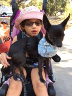 A child is sitting wearing a pink hat and glasses. The child has a black dog with a blue patterned bandana on their lap
