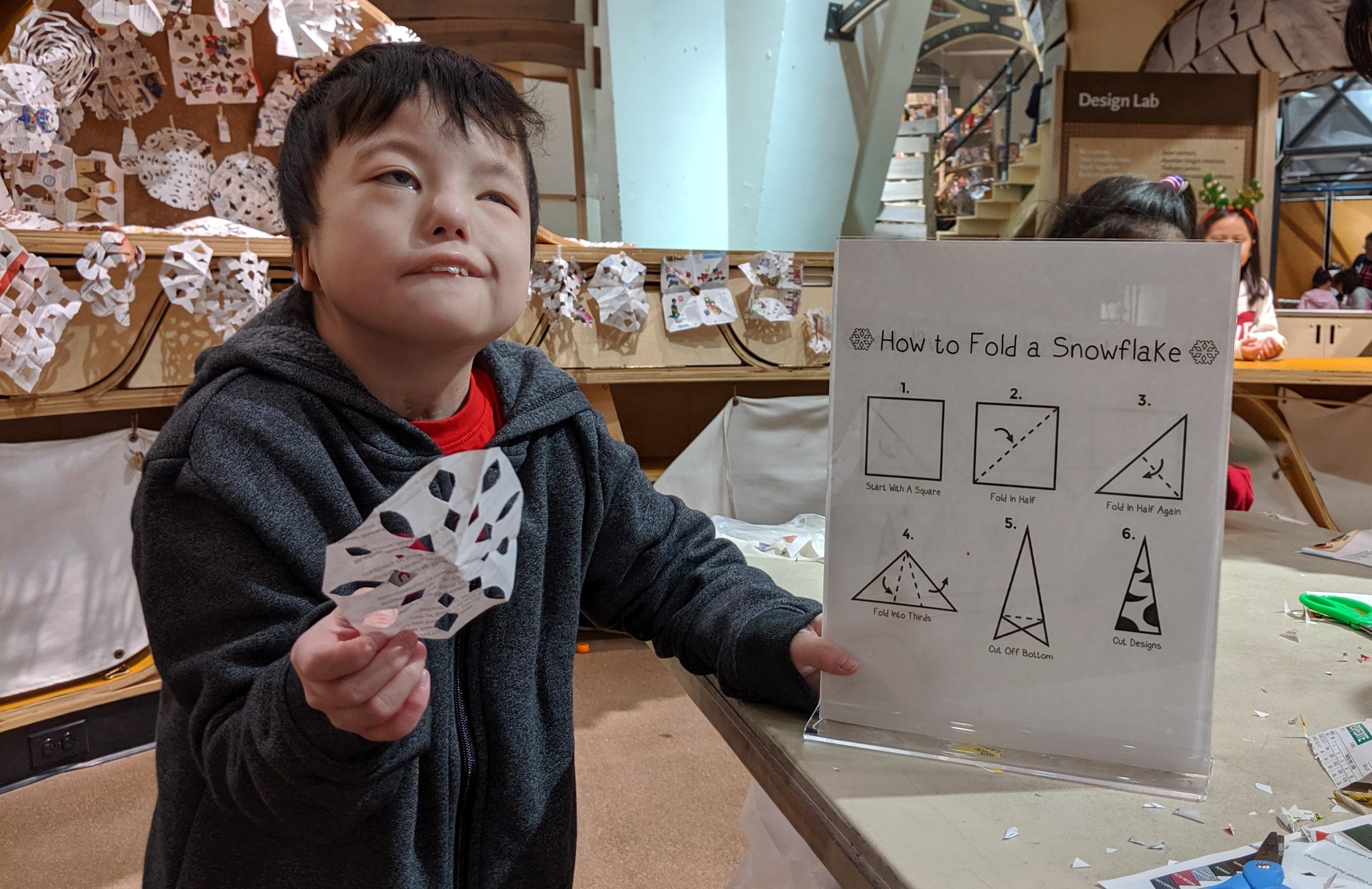 An 11-year-old boy with CHARGE syndrome is holding a paper snowflake in one hand and a paper with illustrations showing how to fold a snowflake in the other.