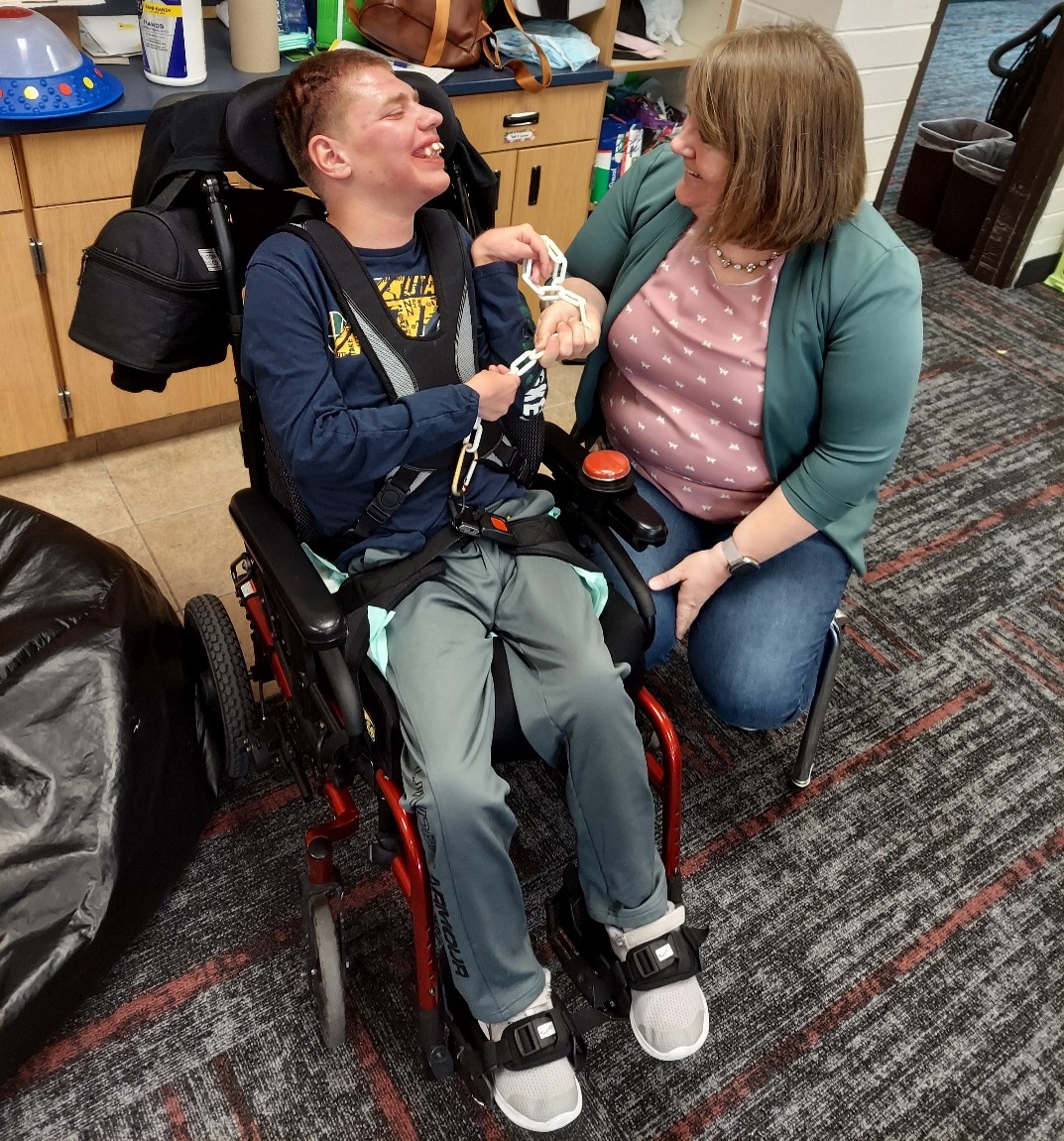 A teacher sits on a low stool by a teenage boy in a wheelchair. They are smiling at each other.