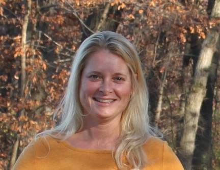 A smiling woman with long blond hair and wearing a yellow sweater is standing in front of trees.