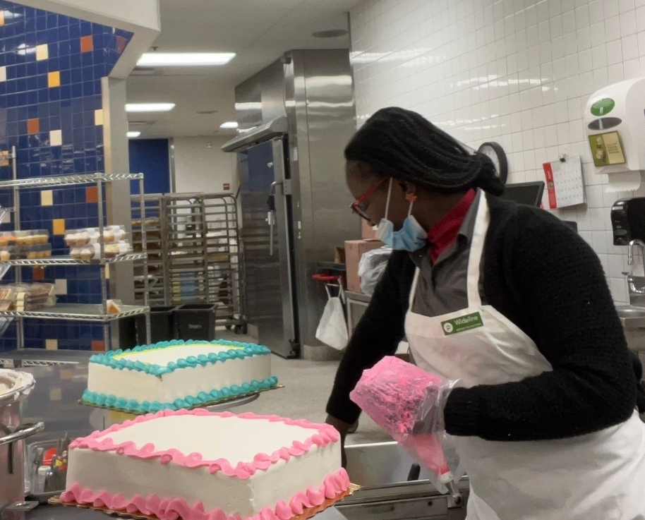 A woman decorates a cake with pink icing in a commercial kitchen.