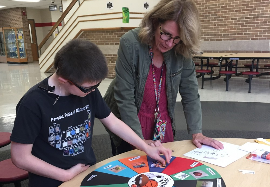 A boy who is deafblind stands at a large desk with his hand on a colorful poster. His intervener stands close by watching him work.