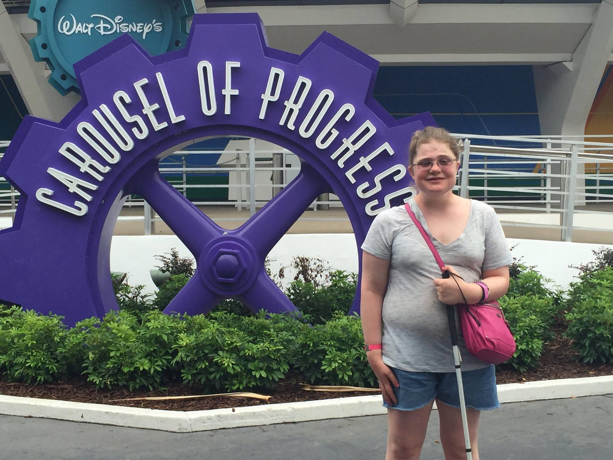 A girl with a grey shirt, shorts, pink bag, and glasses stands in front of a purple gear that says Carousel of Progress and a Walt Disney