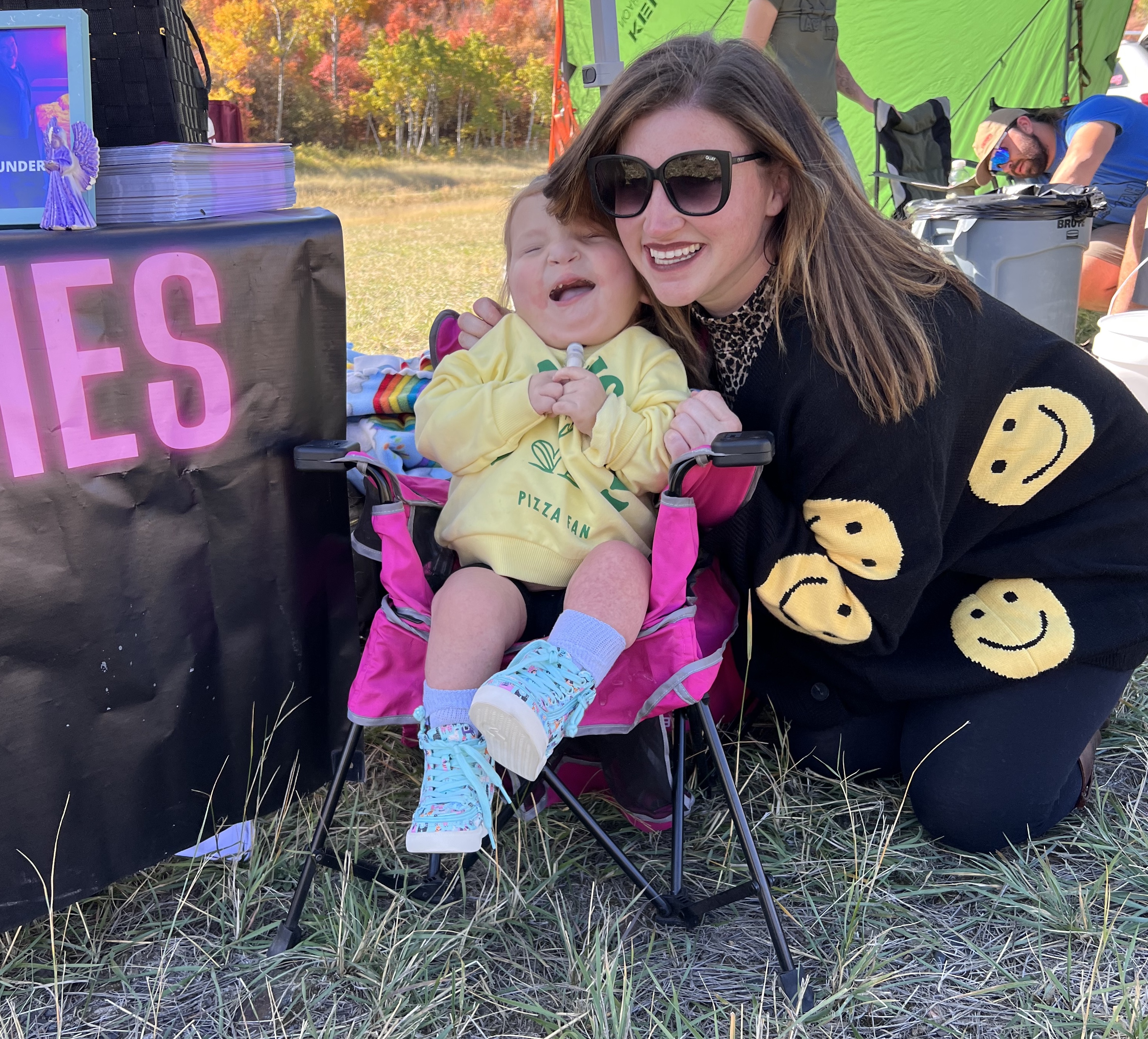 Sloan sits in a chair with her mom standing behind her. They are outside at an event.