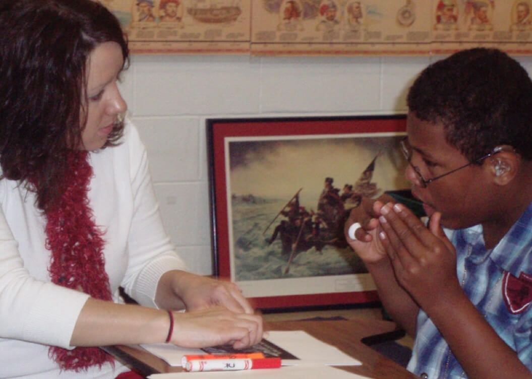 A teacher and a teenage boy with blindness sitting at a table that has papers and markers on it. The boy is signing to the teacher.
