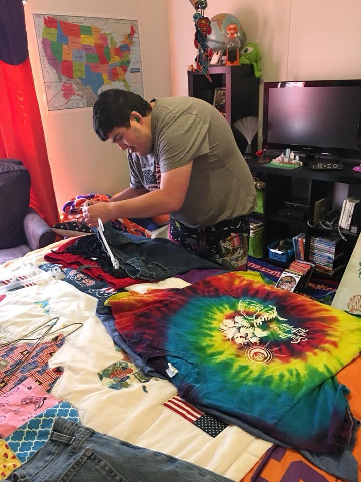 A young man is standing by a bed that contains clothing items. He is attaching a hangar to a pair of pants.