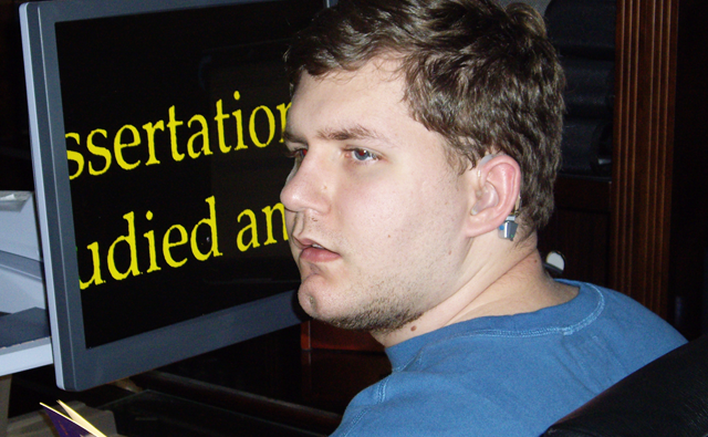 A man with a computer screen behind him that has words in a large yellow font on a black background.