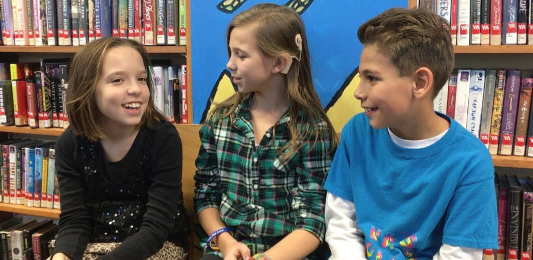 Three students sit next to eachother in front of a library shelf. The student in the middle has a cohlear implant.